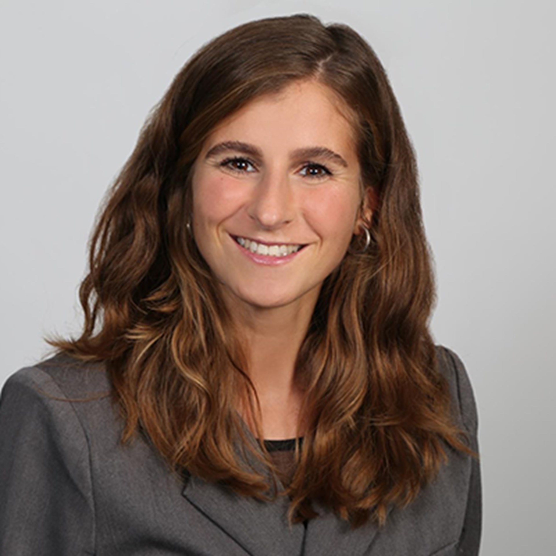 Lächelnde, junge Frau mit langen, brauen Haaren, die eine graue Anzugjacke trägt